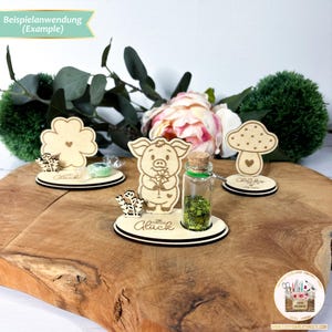 May include: Four wooden figurines, a four-leaf clover, a pig, a mushroom, and a glass bottle filled with green glitter, are displayed on a wooden surface. Each figurine has a small base with the word "Gl&uuml;ck" (German for "luck") written on it.