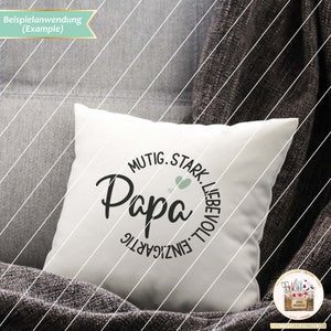 May include: White cushion with a black circle design that says "Papa" in a script font. The circle is surrounded by the words "Mutige, Stark, Liebevoll, Einzigartig" in German.