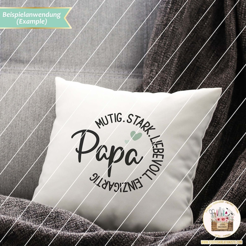 May include: White cushion with a black circle design that says "Papa" in a script font. The circle is surrounded by the words "Mutige, Stark, Liebevoll, Einzigartig" in German.