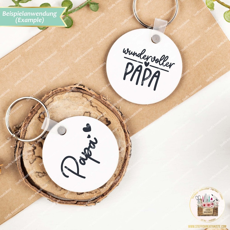 May include: Two white round keychains with black text. One keychain says "wundervoller PAPA" with a small heart above the word "PAPA". The other keychain says "Papa" with a small heart to the left of the word.