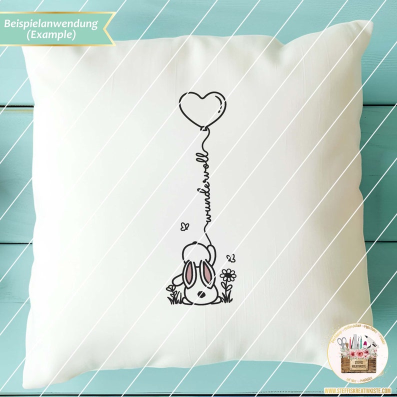 May include: A white pillow with a black line drawing of a bunny holding a heart-shaped balloon. The balloon string spells out the word "wunderwoll".