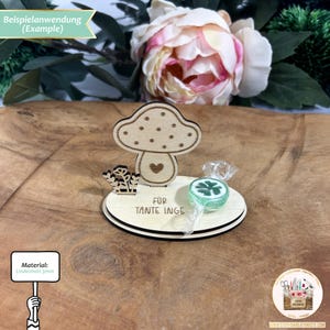 May include: A wooden mushroom-shaped stand with a heart cutout and the text "FOR TANTE INGE" on the base. A green four-leaf clover candy is placed on the stand.