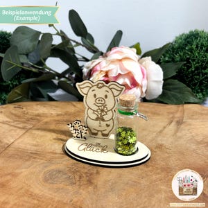 May include: A wooden pig figurine holding a flower, with a small glass jar filled with green glitter. The jar is labeled "Eine Portion Gl&uuml;ck" which translates to "A Portion of Luck".