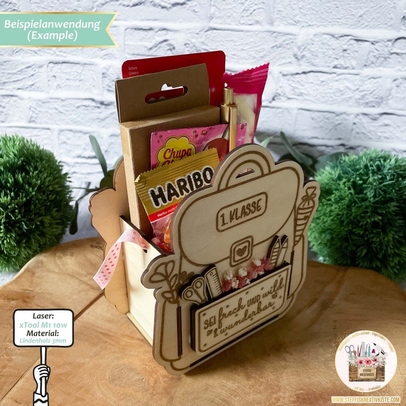 May include: A wooden backpack-shaped box with a 1st grade label, decorated with flowers and the text "SEI frech und wild wunderbar". The box is filled with small gifts, including sweets, pencils, and a pen.