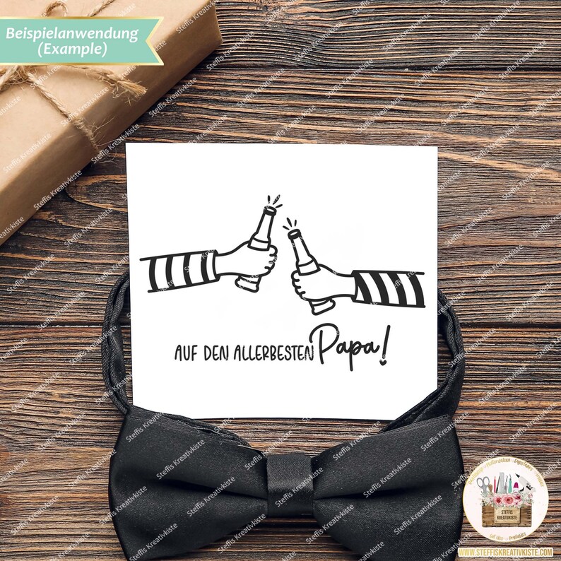 May include: A white card with a black ink illustration of two people clinking beer bottles. The text reads "AUF DEN ALLERBESTEN Papa!"