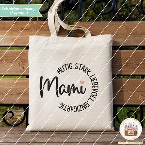 K&ouml;nnte beinhalten: Eine wei&szlig;e Stofftasche mit einem schwarzen Kreisdesign, auf dem "Muti. Stark. Liebevoll. Einzigartig" um das Wort "Mami" mit einem rosa Herzen steht.