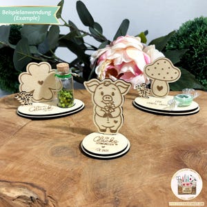 May include: Three wooden decorations with a clover, a pig holding a clover, and a mushroom. Each decoration has a small base with a message. The clover base says "Gl&uuml;ck", the pig base says "Ein Gl&uuml;cks Schweinchen f&uuml;r dich", and the mushroom base says "F&uuml;r Tante Inge".