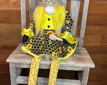 May include: A whimsical bee-themed gnome doll. It features a yellow and black honeycomb patterned dress with sunflower accents, a yellow top, and a straw hat with a golden crown. The doll is seated on a rustic wooden chair and holds a wooden honey dipper.