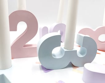 May include: A set of pastel-colored number candle holders. The numbers are 2 and 3, and they are made of wood. The candle holders are designed to hold standard-sized candles.