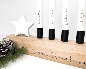 May include: A wooden Advent candle holder with four white candles, each with a black base and a number written on it. The numbers are "eins", "zwei", "drei", and "vier". The holder has the words "Merry Christmas" engraved on it. A white star sits on top of the holder.