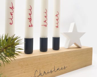 May include: A wooden Advent candle holder with four white candles, each marked with a red number. The holder has a white star and the words "Merry Christmas" are written on the front. The candles have black bases.