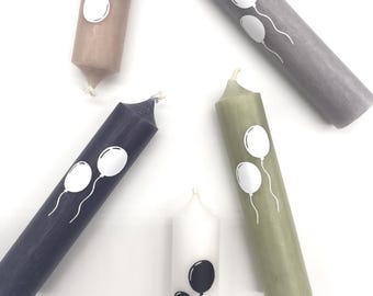 May include: A collection of pillar candles in various colors, including gray, navy, and olive green. Each candle features a white balloon design. One white candle has a black balloon design. The candles are arranged on a white surface.