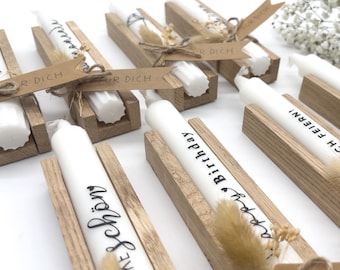 May include: White candles with black lettering, displayed in wooden holders. Some candles have the words "Happy Birthday" or "Schön" written on them. The holders are decorated with dried flowers and small paper tags.