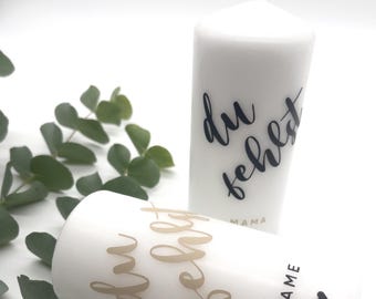 May include: Two white pillar candles with the text "du fehlst" in black and gold lettering. The candle on the left has the text in gold and the candle on the right has the text in black. The candle on the right also has the word "mama" in black lettering below the text. The candle on the right has the word "NAME" in black lettering and a small black heart below the text.