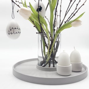 Könnte beinhalten: Ein graues Tablett mit einer durchsichtigen Glasvase, gefüllt mit weißen Tulpen und Zweigen. Zwei weiße, eiförmige Kerzen stehen in grauen Haltern auf dem Tablett. An der Vase hängt ein weißes Ei mit dem Schriftzug "Frohe Ostern".