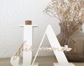 May include: White wooden decorative letters spelling "JA" with the word "für immer" in cursive. The "J" has a cork stopper. A vase with dried flowers is in the background.