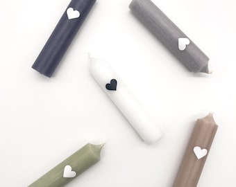 May include: Five candles with different colored waxes and heart-shaped stickers. The candles are white, gray, light brown, dark gray, and green.