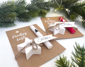 May include: Christmas-themed gift set featuring a white star-shaped ornament tied with twine, a white candle with a black tree design, and a brown card with the text "frohes fest". A red candle is also included.