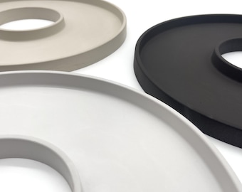 May include: Three modern, circular serving trays in neutral colors. The trays are in shades of white, gray, and black. Each tray has a raised, circular center. The trays are made of a smooth, matte material.