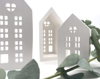 May include: Three white ceramic houses with cut-out windows and heart-shaped details. The houses are arranged in a row, with the largest house in the back and the smallest house in the front. The houses are sitting on a white surface with green leaves in the foreground.