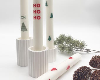 May include: Three white candles with Christmas-themed designs. The candles are in white holders with a ribbed texture. The candles are decorated with green Christmas trees and the words "HO HO HO" in red. There are also pine branches and pine cones in the image.