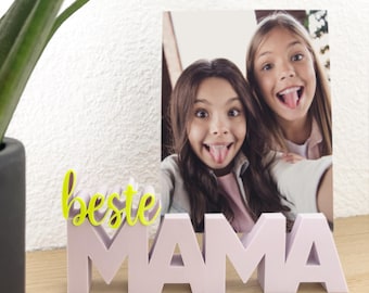 May include: A pink "MAMA" block photo holder with a photo of two girls sticking out their tongues. The word "beste" in yellow script is in front of the photo holder. A green plant is in a black pot to the left.