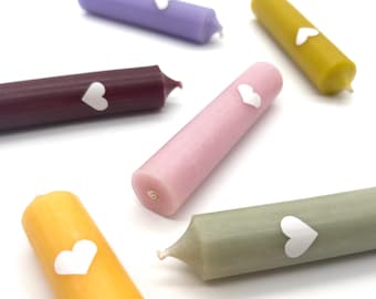 May include: Six colorful candles with white heart stickers on them. The candles are arranged in a semi-circle on a white surface.
