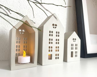 May include: Three white ceramic houses with cut-out windows and a heart shape at the top. The houses are lit from inside with small LED tea lights.
