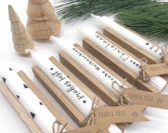 May include: White candles with black text in wooden holders, some with Christmas greetings. The candles are decorated with small black tree designs. Wooden Christmas tree figurines and greenery are in the background.