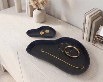 May include: Two matte dark charcoal, organic-shaped trays with raised edges. The smaller tray holds two gold-toned circular earrings, while the larger tray contains a thin gold chain necklace and two gold-toned bangles of varying thicknesses.