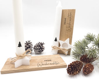 May include: A Christmas-themed display featuring white candles with black tree graphics, wooden candle holders, and pine cones. The wooden holders have the words "Frohe Weihnachten" engraved. A star-shaped candle holder is tied with twine.