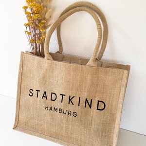 Op de afbeelding: Een bruine jute tas met zwarte handvatten en de tekst "STADTKIND HAMBURG" gedrukt op de voorkant.
