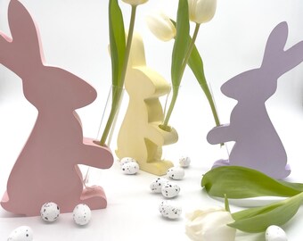 May include: Three wooden bunny shaped vases in pink, yellow, and purple, each holding a single white tulip. The bunnies are sitting on a white surface with scattered white speckled Easter eggs.