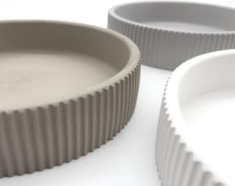 May include: Three round concrete bowls with a ribbed edge. The bowls are in shades of gray, beige, and white.