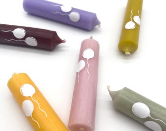 May include: Six colorful birthday candles with white balloon designs. The candles are in shades of purple, maroon, pink, yellow, and green.