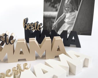 May include: Wooden block letters spelling "MAMA" in beige, black, and white, with the word "beste" in gold script. A black and white photo of a child is in the background. The letters are arranged on a white surface.
