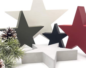 May include: A collection of decorative wooden stars in various colors, including white, black, red, and gray. The stars are arranged on a white surface, with a pinecone and evergreen sprig in the foreground, suggesting a holiday theme.