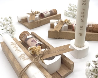 May include: Three wooden candle holders with a white candle, a small glass vial with a rolled up bill, and a small glass vial with a rolled up bill and a small white heart. The candle holders have a small tag that says "Alles Liebe" and the candle has the date "15. März 2021" engraved on it.