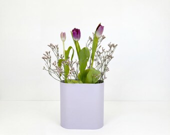 May include: A light lavender oval-shaped vase containing a floral arrangement of three tulips in shades of purple and pink, accompanied by delicate sprigs of small, light-colored filler flowers with thin green stems and broad green leaves.