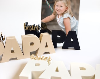 May include: Wooden block letters spelling "PAPA" in various colors, including black, beige, and white. Some blocks have the word "bester" in gold script. A framed photo of a young girl is in the background.