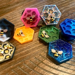 May include: Assortment of hexagonal phone grips with a honeycomb design and a bee accent. The grips are made of resin and come in a variety of colors, including pink, orange, green, blue, and black.