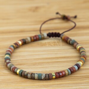 May include: A multi-colored beaded bracelet with a brown cord closure. The beads are made of natural stone and have a variety of colors, including red, brown, green, and gold. The bracelet is adjustable and can be worn by both men and women.