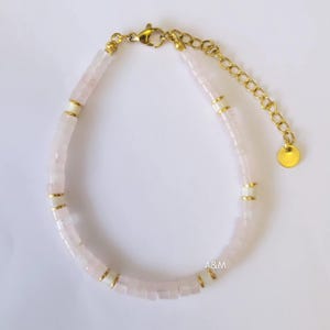 May include: A delicate bracelet featuring small, square, light pink beads interspersed with white and gold-colored accents. The bracelet has a gold-colored chain and clasp. The letters "A&M" are visible on the bottom right.