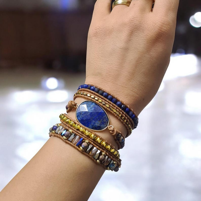 May include: A layered bracelet with a large oval lapis lazuli gemstone charm. The bracelet is made with brown leather cord, gold beads, and blue and gold glass beads.