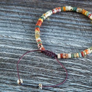May include: A multi-colored beaded bracelet with a brown cord closure. The beads are made of various shades of brown, red, green, and white.
