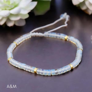 May include: A delicate bracelet made with small, iridescent white beads and gold accents. The bracelet is strung on a white cord and has a sliding knot closure.
