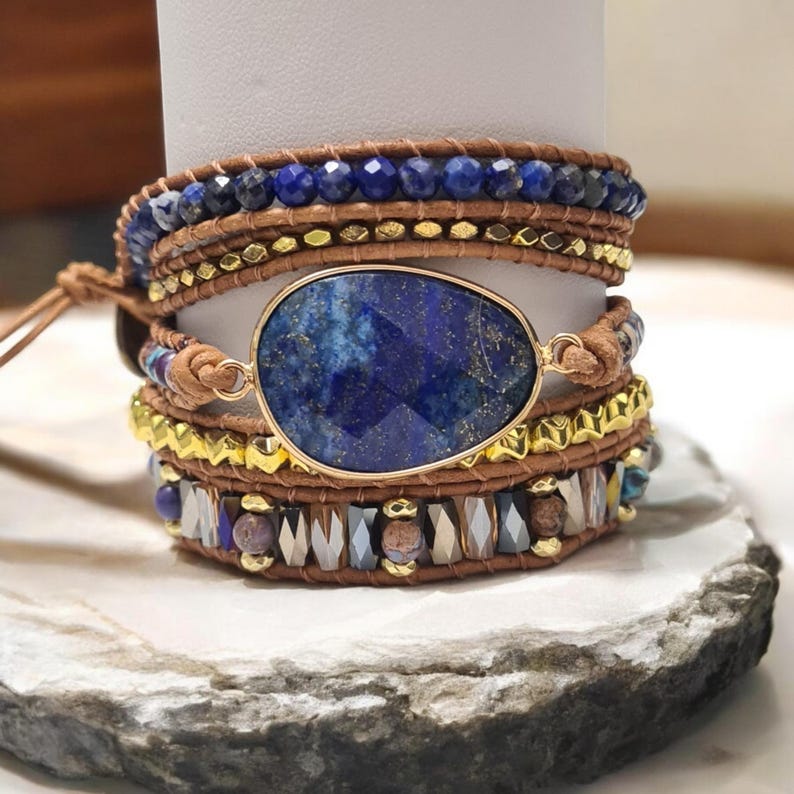 May include: A multi-strand wrap bracelet with a large, oval-shaped lapis lazuli stone. The bracelet is made with brown leather cord, gold beads, and blue and black beads.