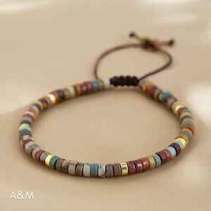 Könnte beinhalten: Ein mehrfarbiges Armband mit braunen, roten, goldenen und grünen Perlen. Das Armband ist aus einem braunen Band gefertigt und hat einen Knotenverschluss.