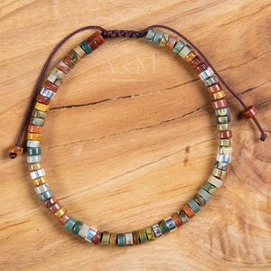 May include: A multi-colored stone beaded bracelet with a brown cord closure. The beads are small and round, and they are arranged in a random pattern. The bracelet is on a wooden surface.