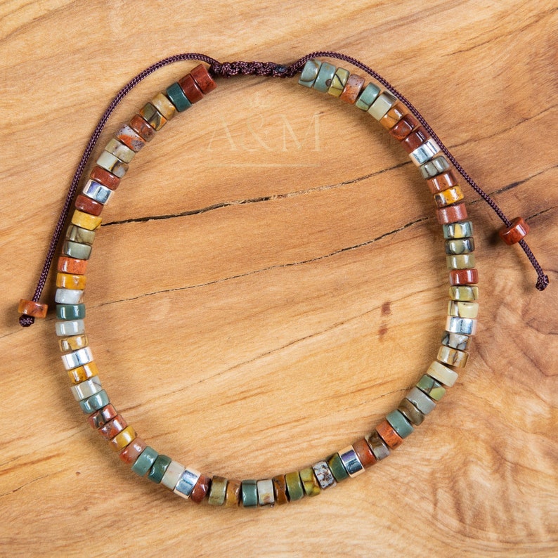 May include: A multi-colored stone beaded bracelet with a brown cord closure. The beads are small and round, and they are arranged in a random pattern. The bracelet is on a wooden surface.